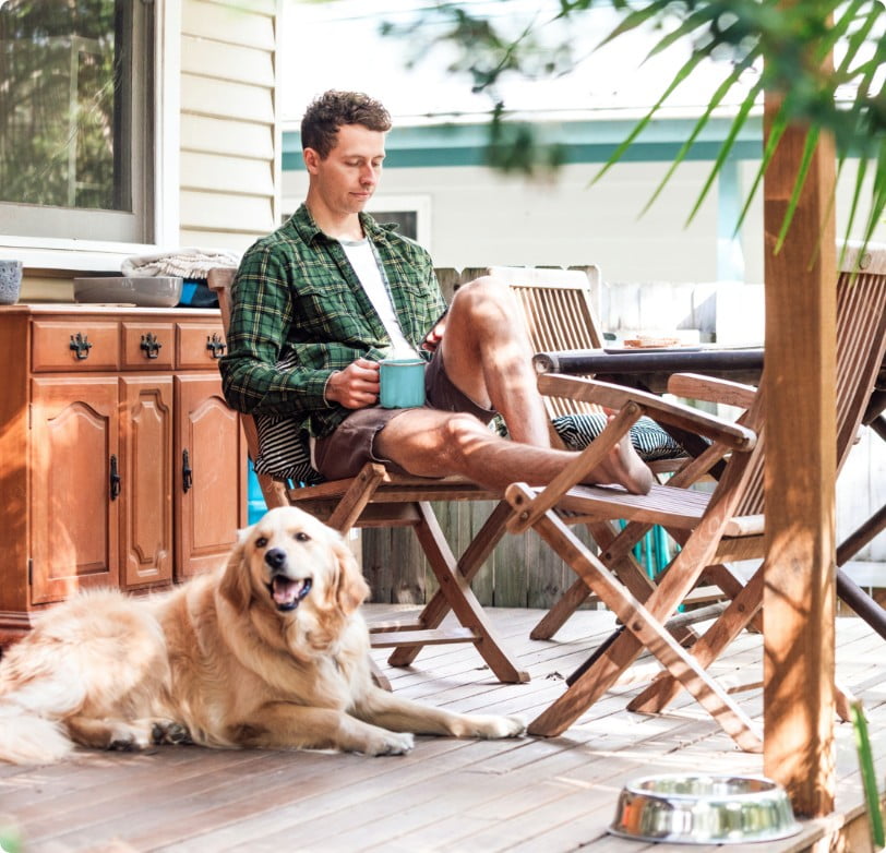 Man sitting outside his home on a rustic chair, researching home buying on a phone. A Golden Retriever is sitting at his feet.